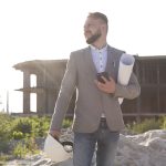 Mężczyzna w eleganckiej marynarce, trzymający zwinięte plany budowlane i telefon, stoi na piaszczystym placu budowy z betonowym szkieletem budynku w tle. Może to być prawnik budowlany Łódź podczas wizytacji inwestycji.